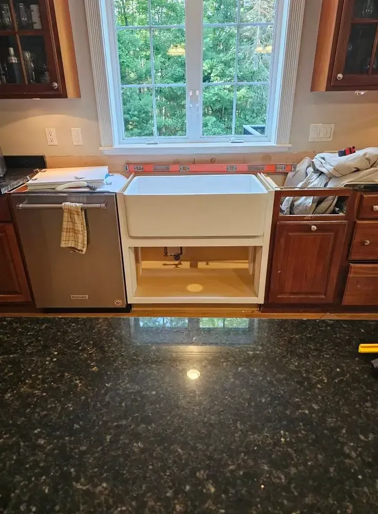 Kitchen with installed farmhouse sink, cabinets, dishwasher, and window overlooking trees.
