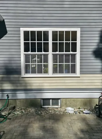 White framed window on gray and beige siding. Small basement window below. Brick patio and landscaping.