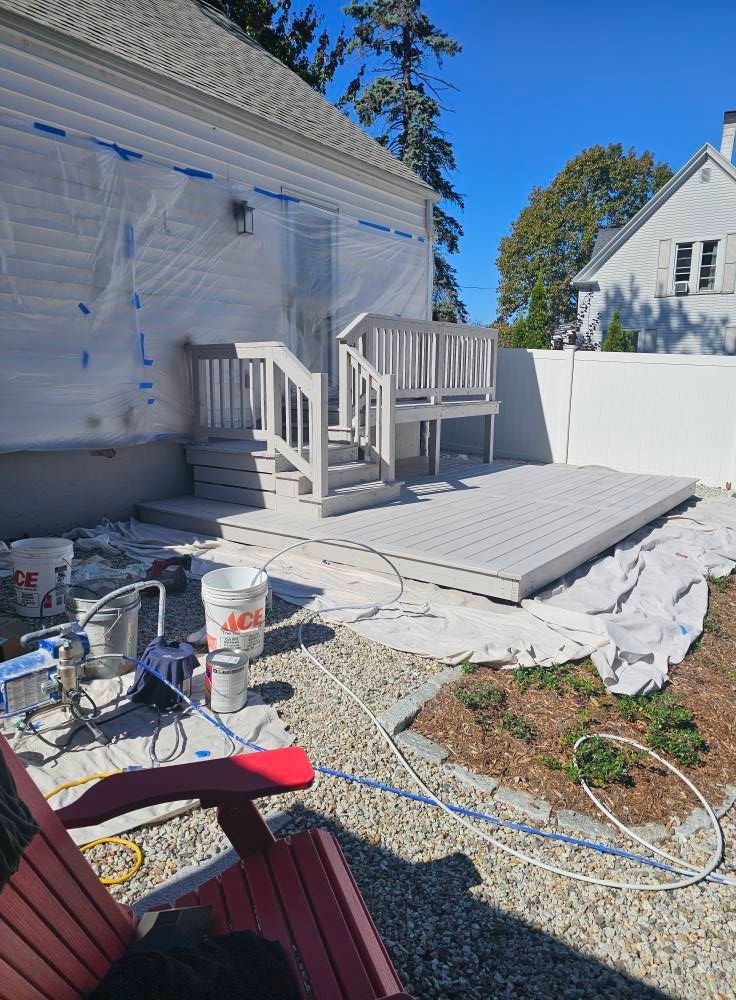 A deck and house being painted light gray outdoors. Painter's equipment and red chair are visible.