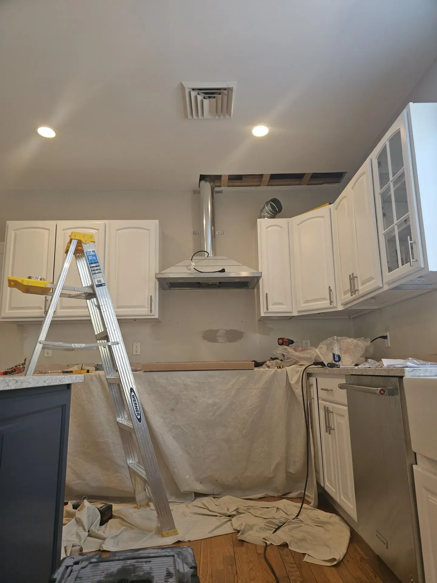 A kitchen undergoing renovation with white cabinets, a range hood, and a ladder placed in the center of the room.