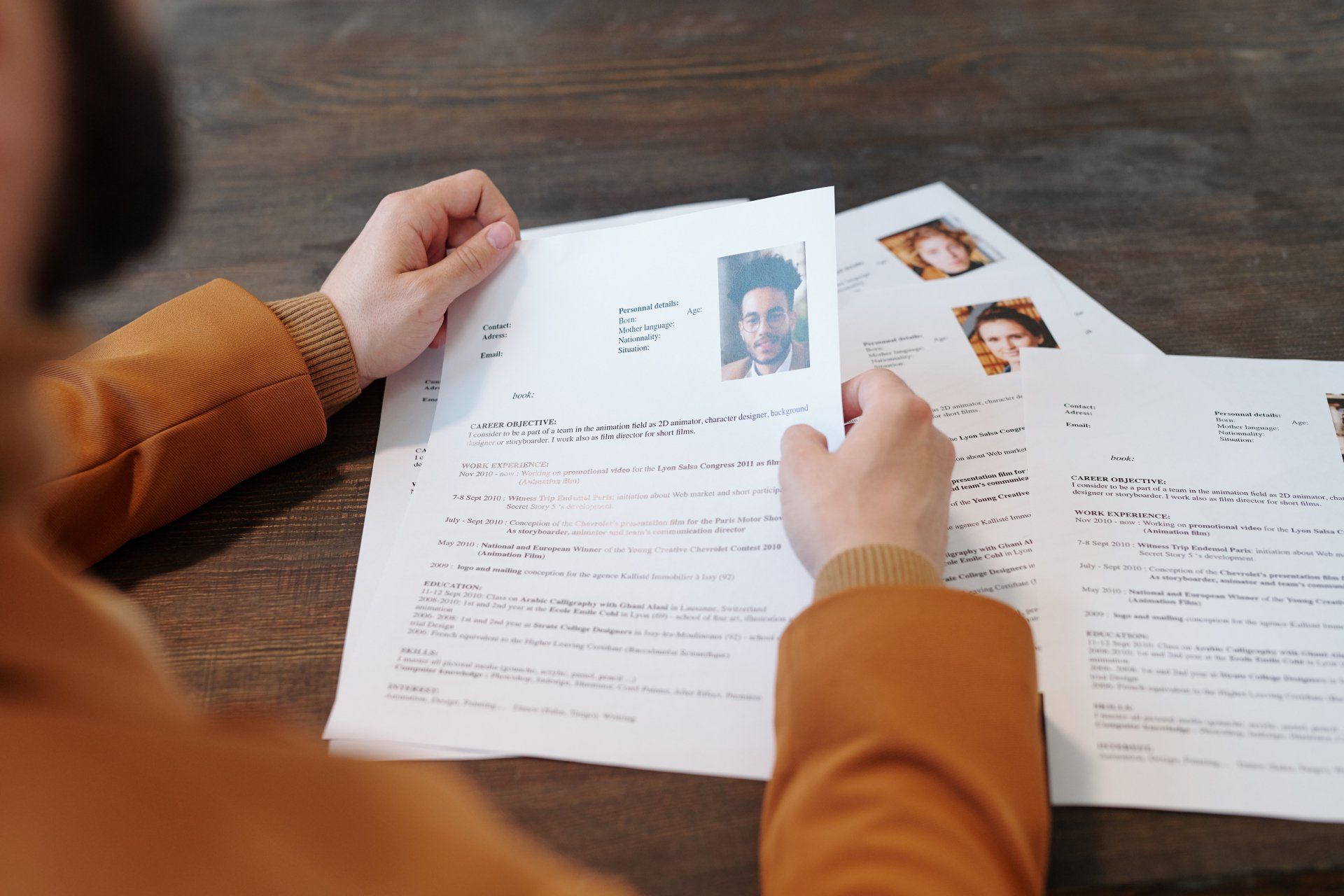 An image looking over the shoulder of an HR recruiter wearing an orange jacket as she sifts through a pile of resumés.