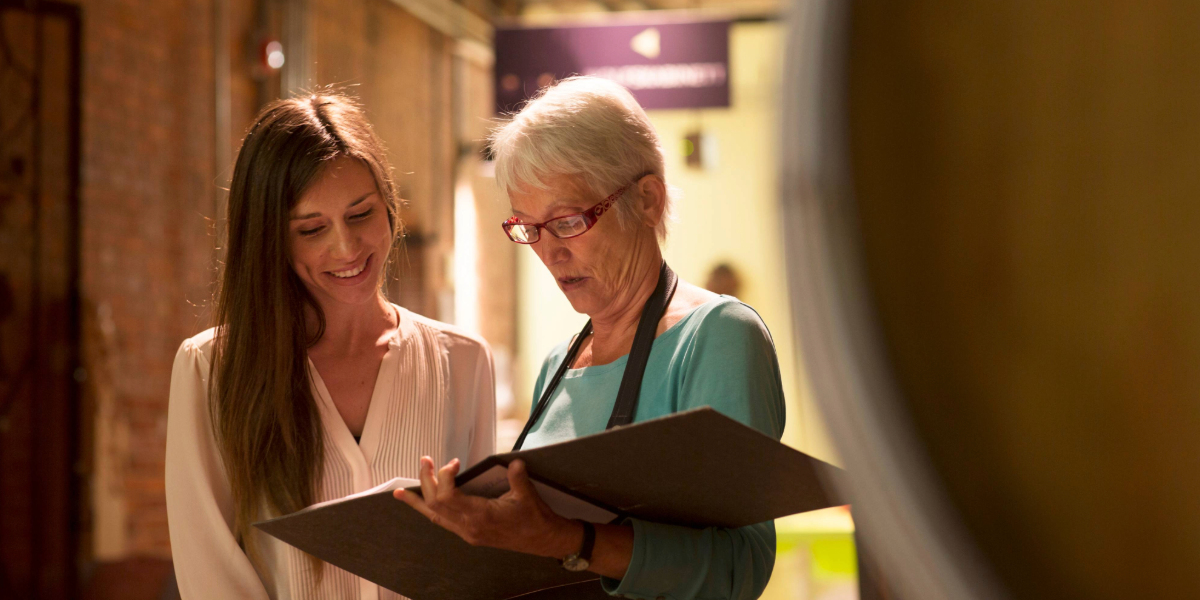 Two women working in a distillery and looking at a log book 