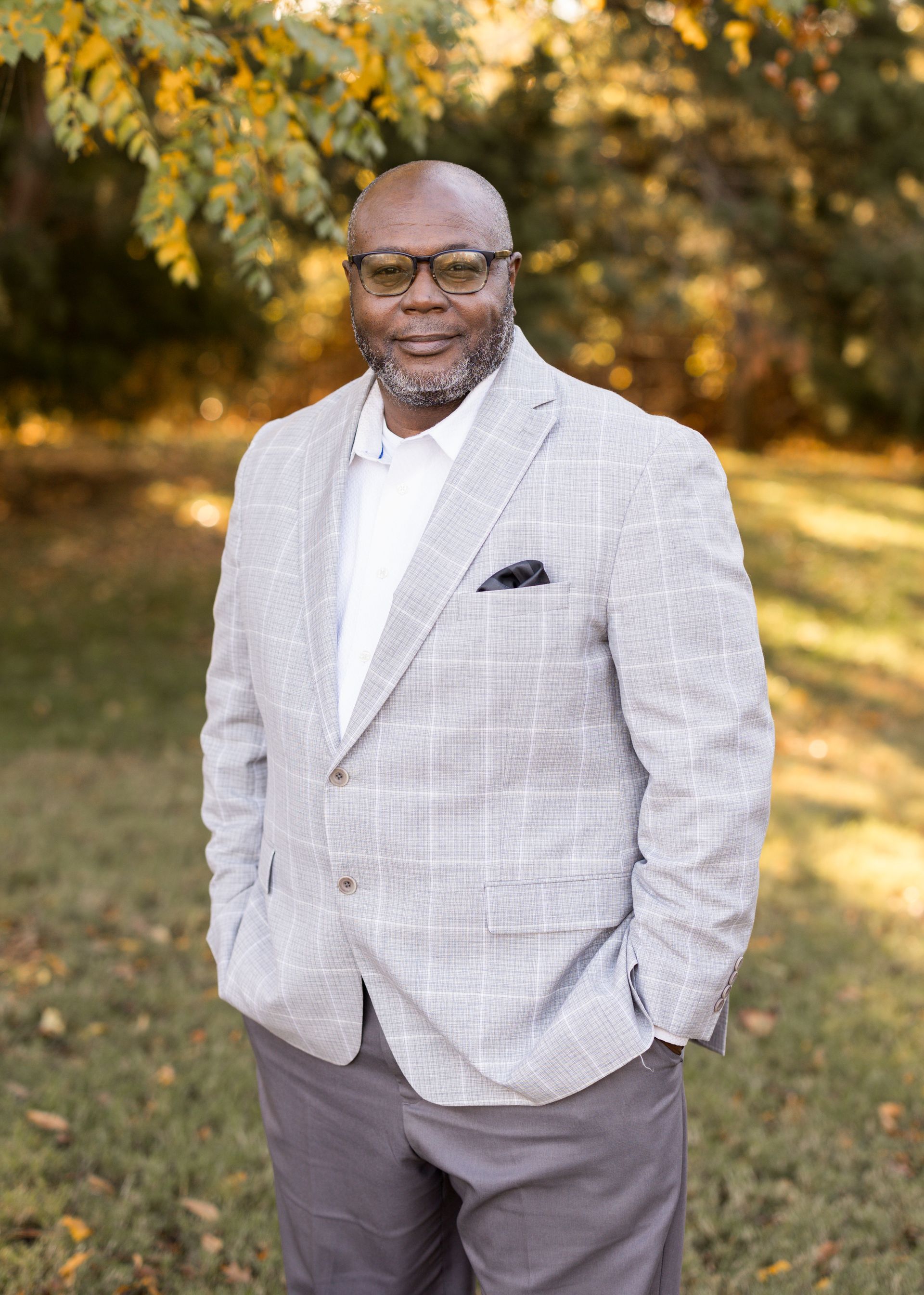 A man in a suit and glasses is standing in a field with his hands in his pockets.