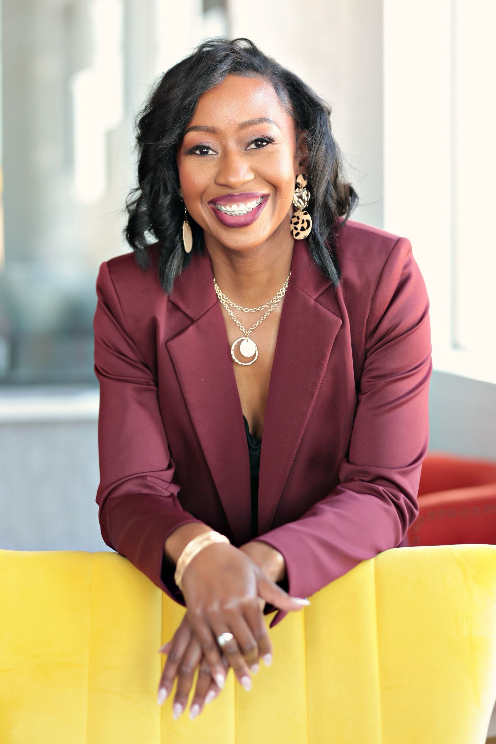 A woman in a maroon jacket and necklace is sitting on a yellow chair.