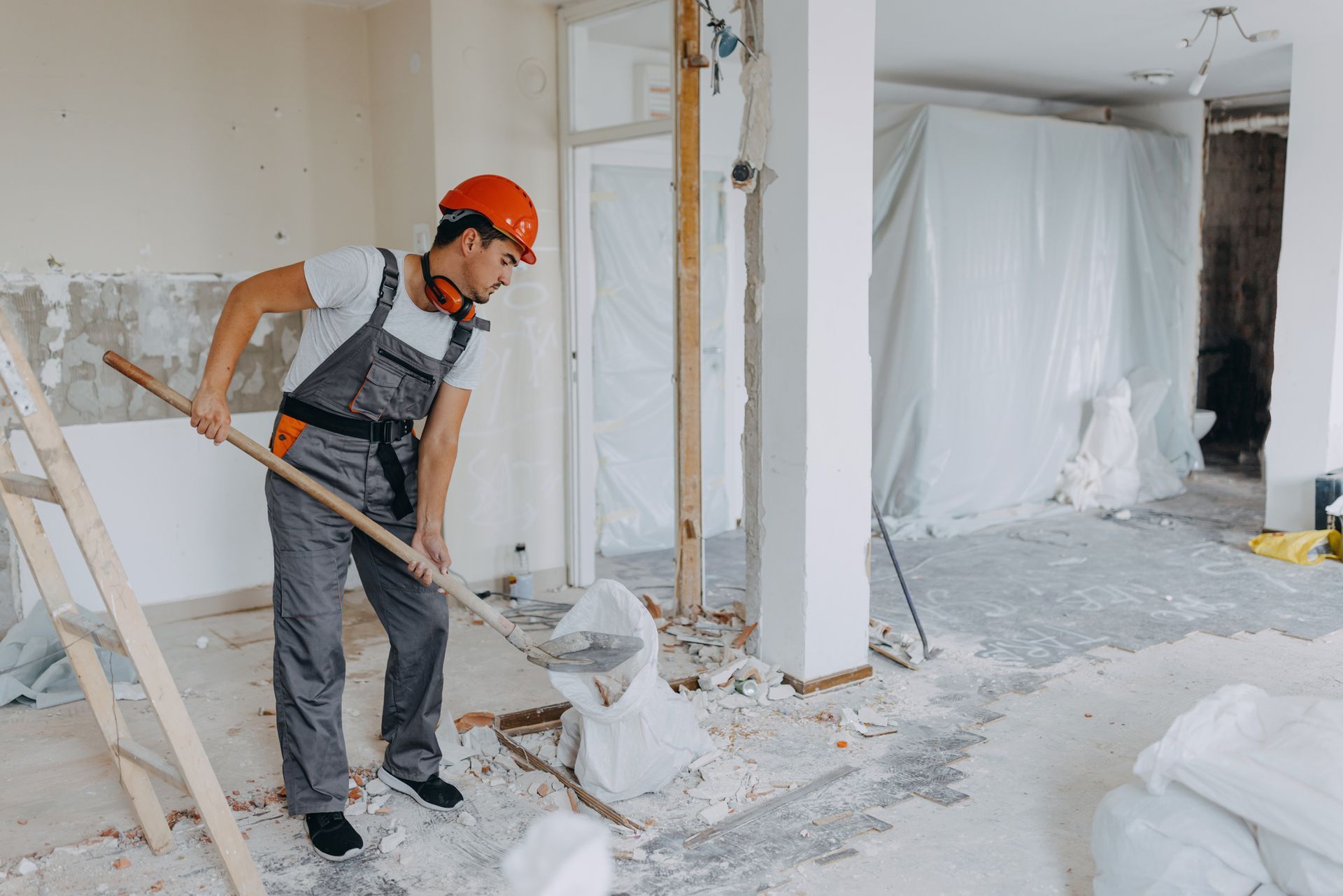 Construction worker in orange helmet shovels debris in a room during renovation.