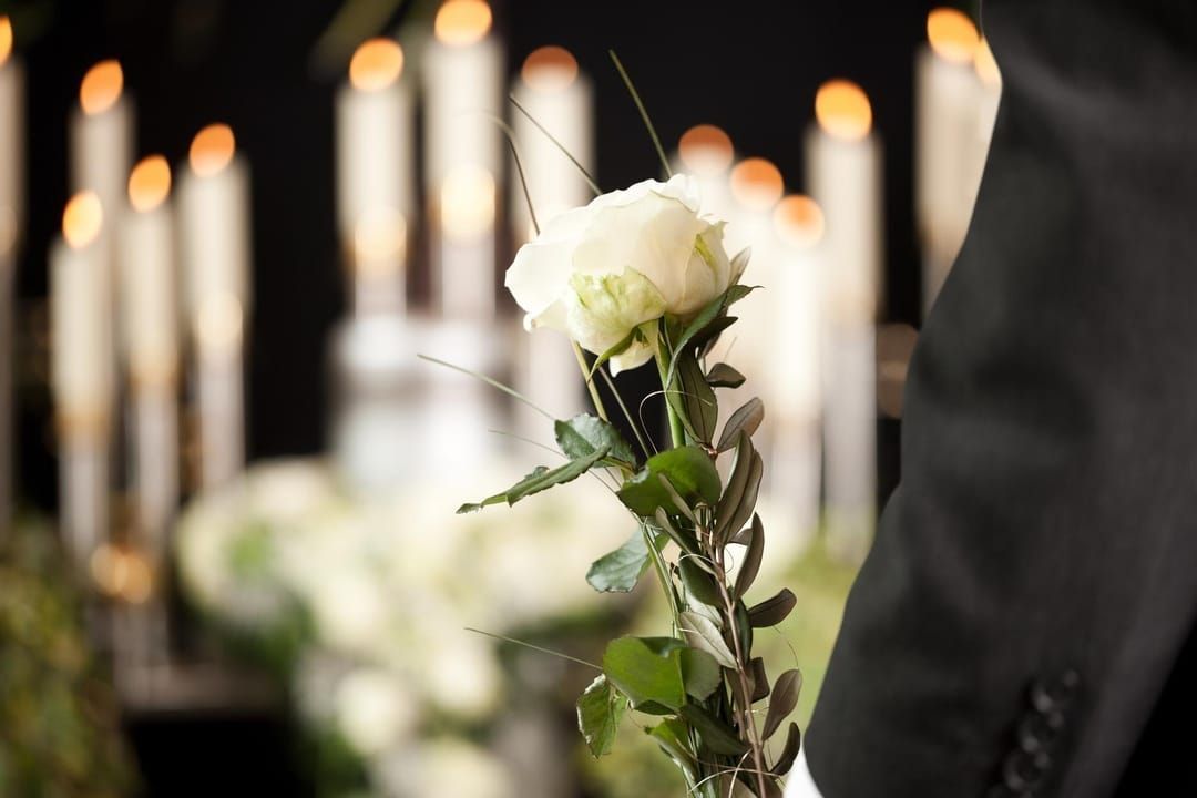 A person in a dark suit holds a single white rose in front of a background of blurred, glowing candles.