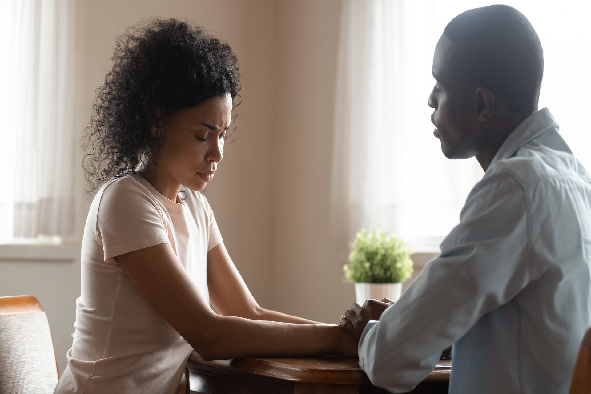 Two people sitting at a wooden table, holding hands with their heads bowed in a quiet, solemn moment.