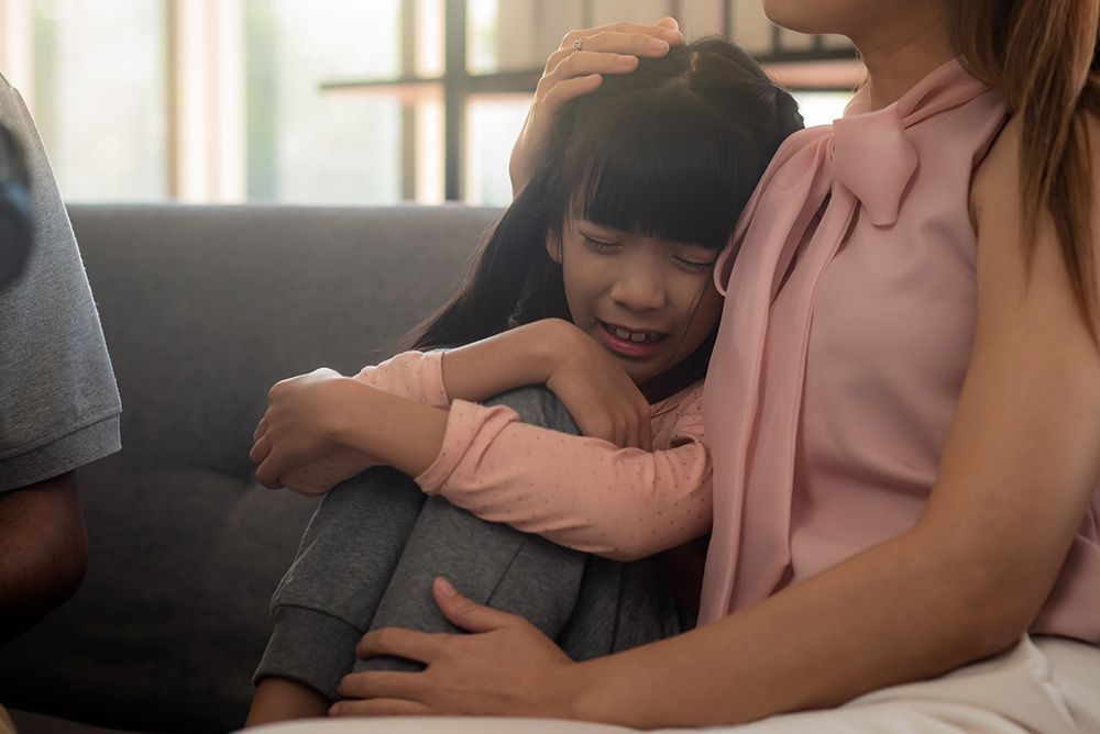 A person in a pink shirt comforts a young child who is curled up on a sofa, looking upset or distressed.