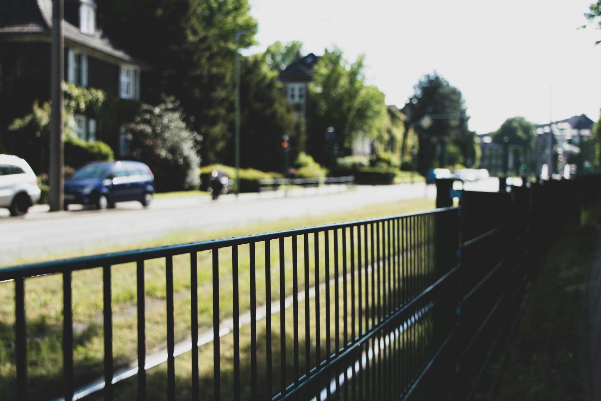 Street view with cars, houses, trees, and a black fence in the foreground