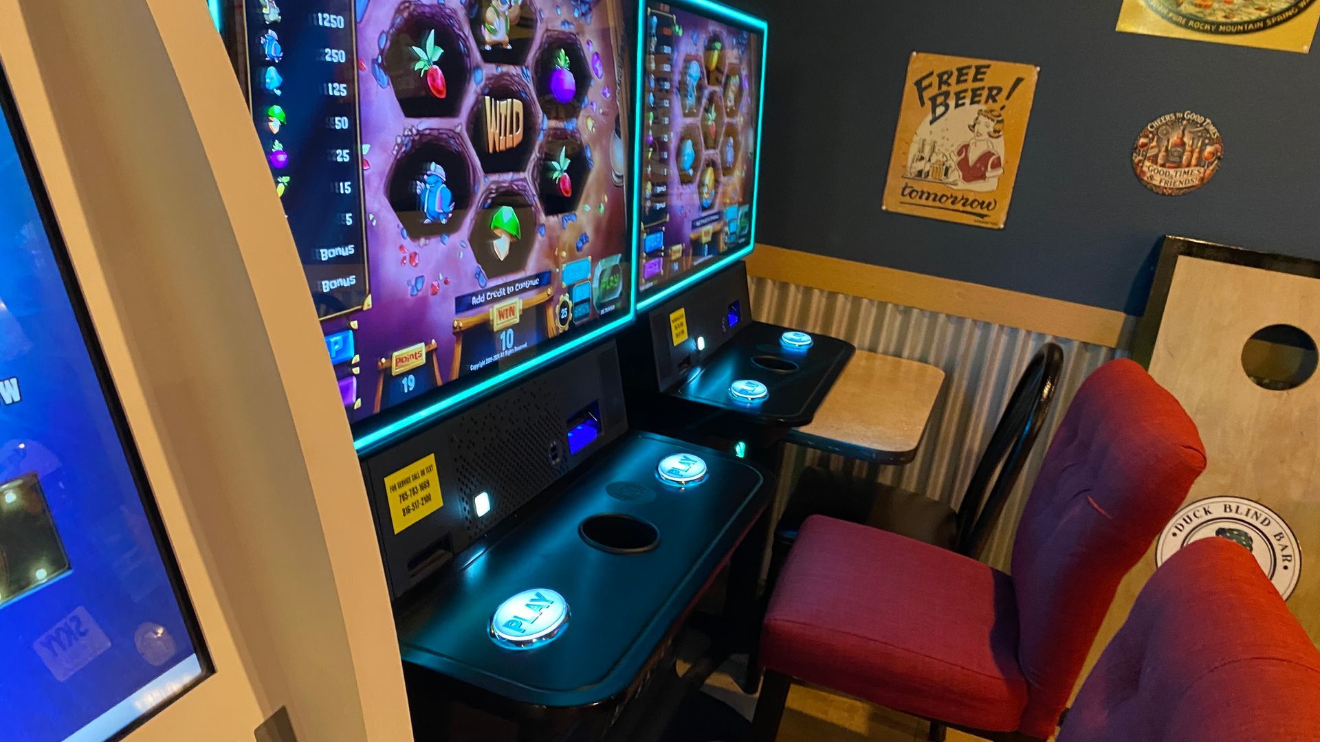 Gaming area with two colorful slot machines, a small table, and red chairs in a bar setting.