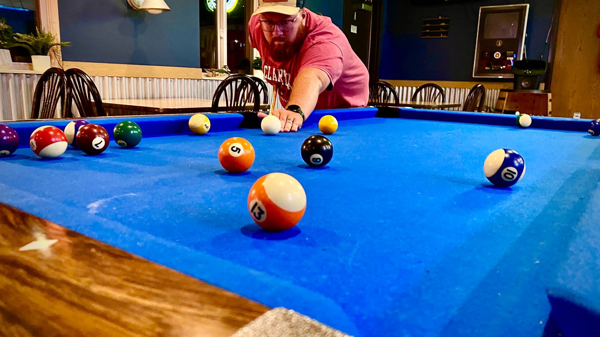 Man in red shirt playing pool at a table with various colored balls in a bar setting.