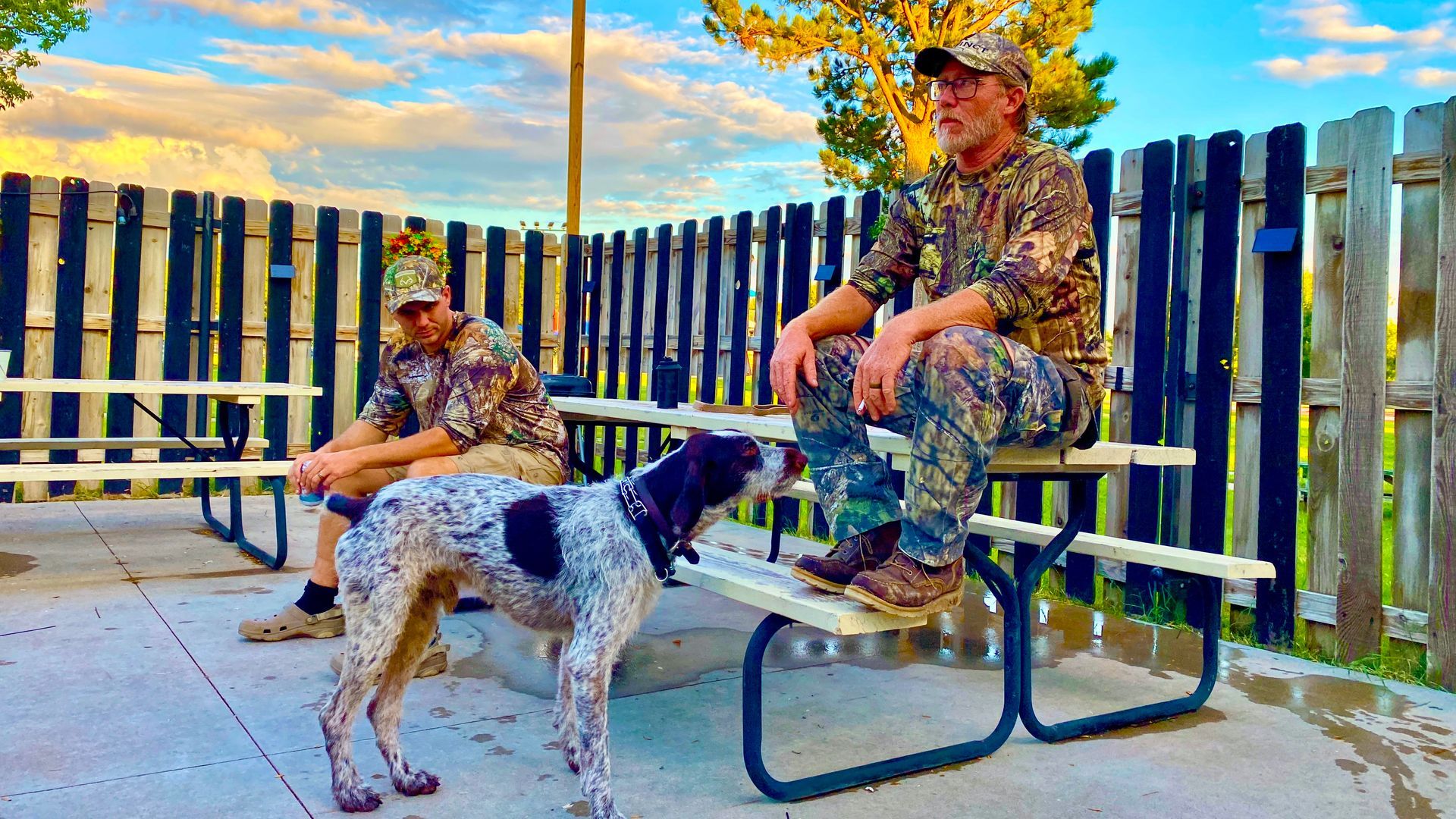 Two men in camouflage with a spotted dog, seated at picnic tables outdoors near a fence.
