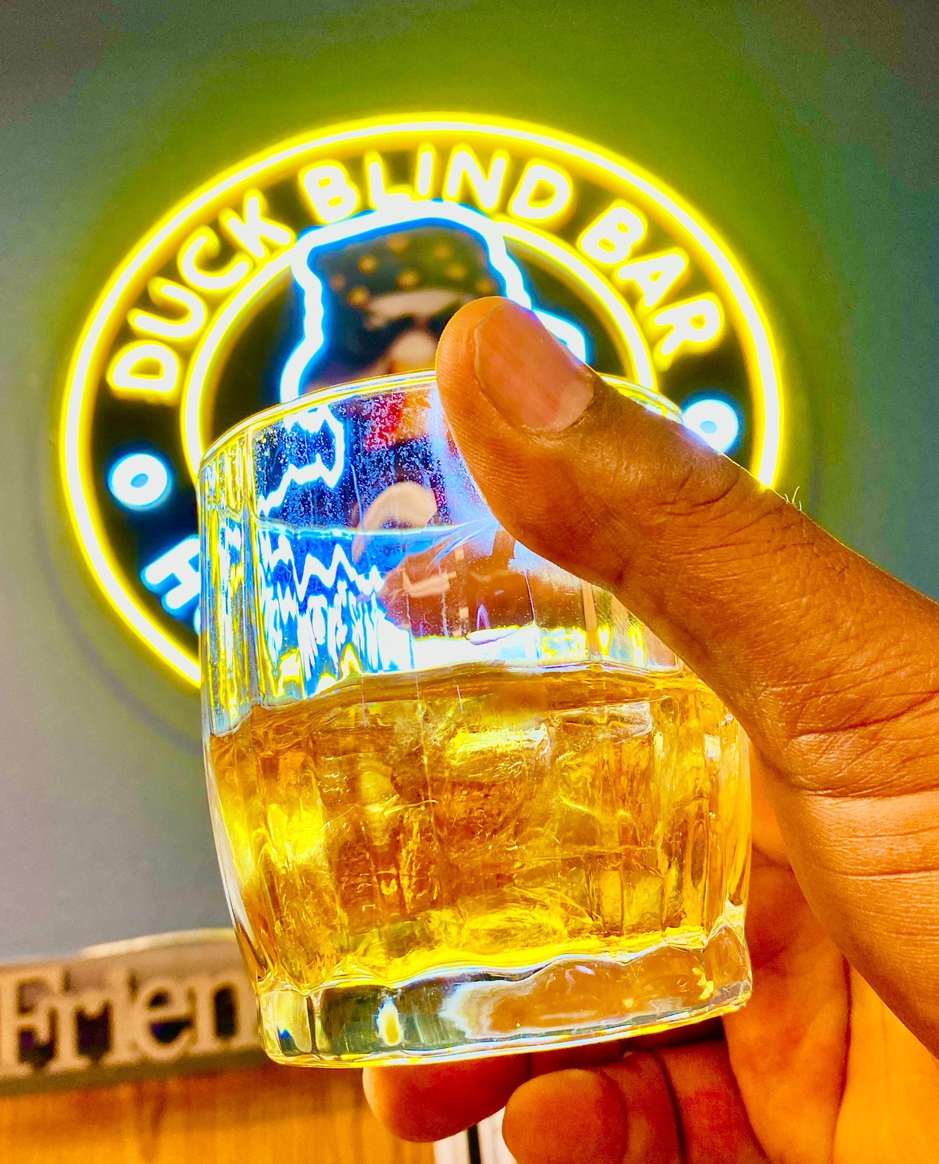 Hand holding a shot glass of amber liquid with the Duck Blind Bar neon sign in the blurry background.