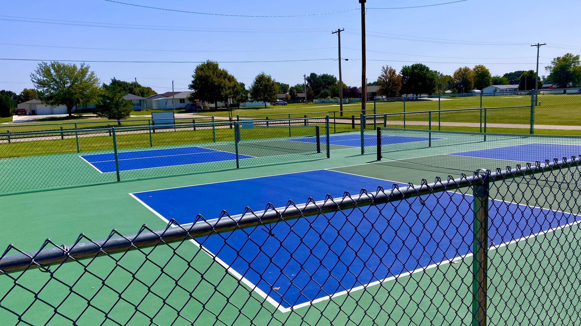 Pickleball courts with blue and green surfaces, fenced in, with poles and a grassy background.