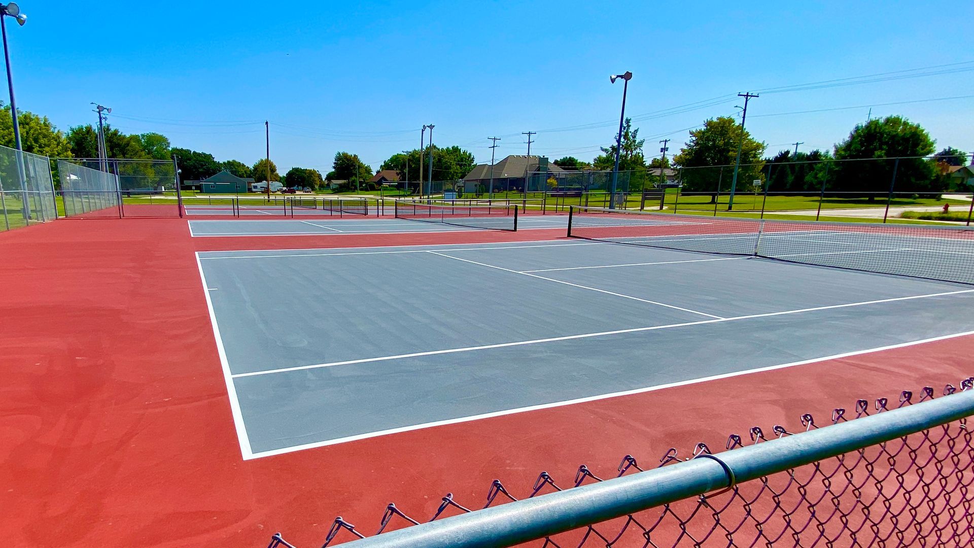 Outdoor tennis courts, red and gray surface, net, chain-link fence, blue sky.