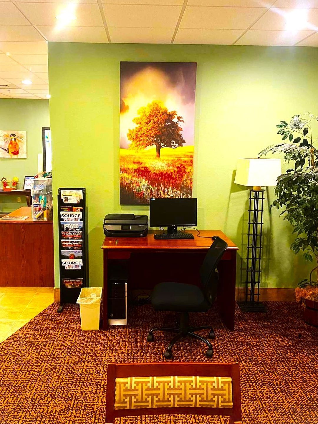 Desk with computer, printer, and tree art on a green wall in an office. Magazine rack and plant on either side.