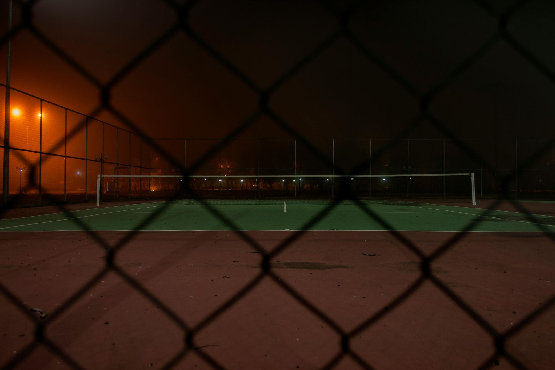 Tennis court at night, viewed through a chain-link fence. Court is lit, with the net visible.