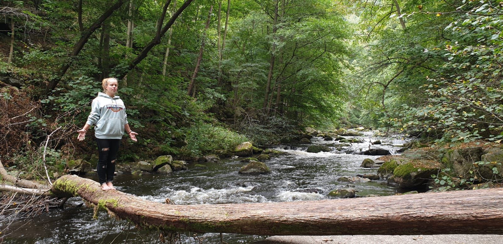 Frau steht in Berghaltung auf einem Baumstamm über einem Fluss im Wald