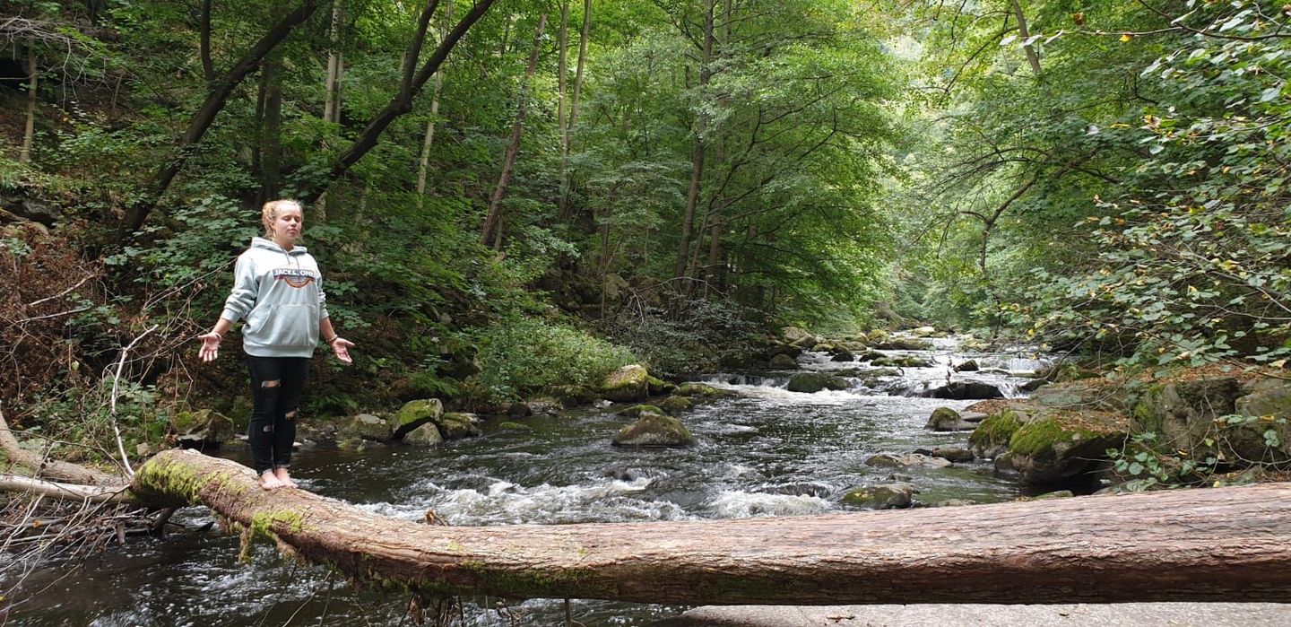 Alena Thielert im Wald: Stehend auf einem Baumstamm in aufrechter, meditativer Yoga-Pose, Augen geschlossen, umgeben von Bäumen

