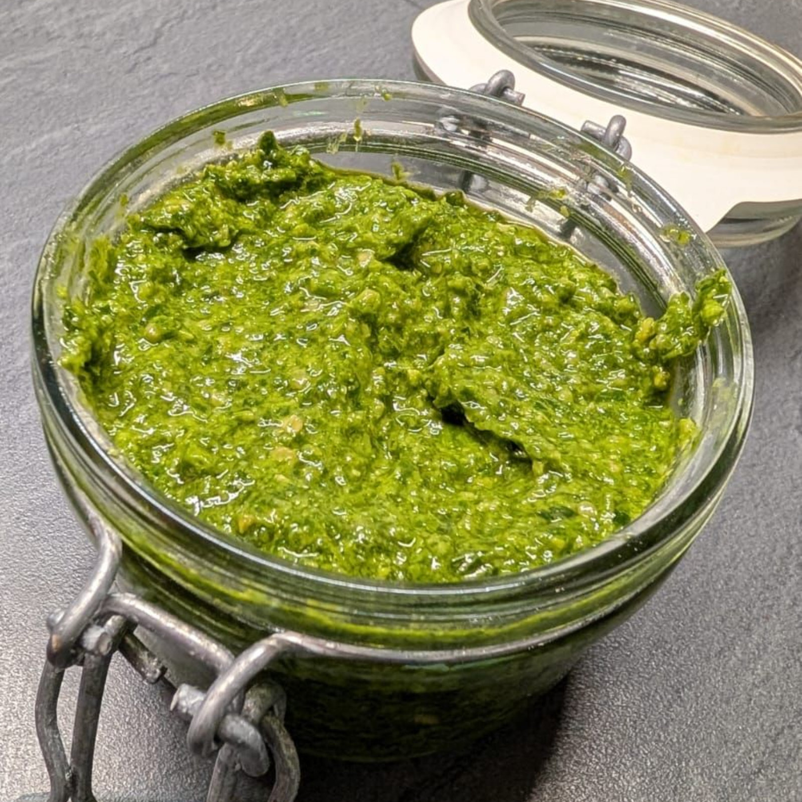 A small glass jar filled with vibrant green pesto sauce, sitting on a dark grey surface.
