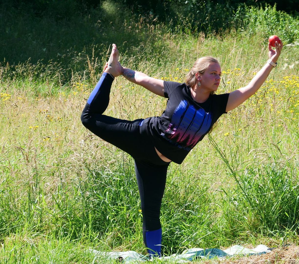 Eine Person balanciert in einer Yoga-Pose auf einem Bein auf einer Wiese und hält einen roten Ball in der Hand.