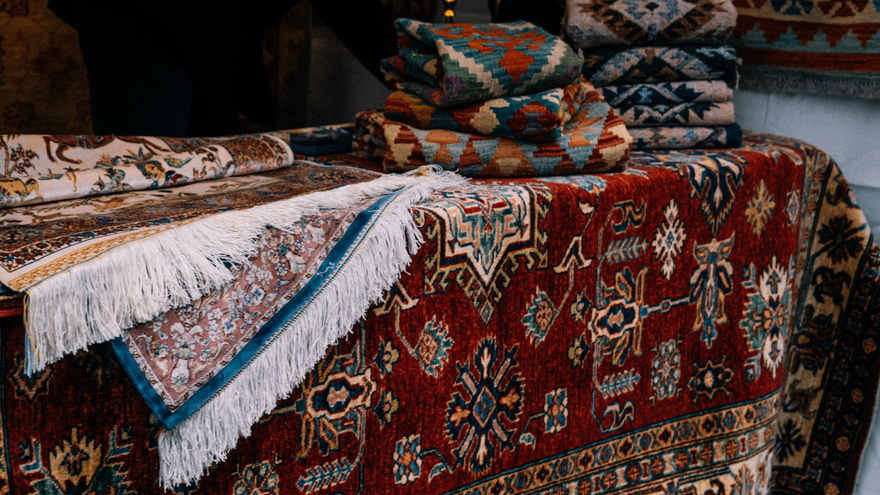 Stacked patterned rugs and cushions in rich red, blue, and beige on a textile display
