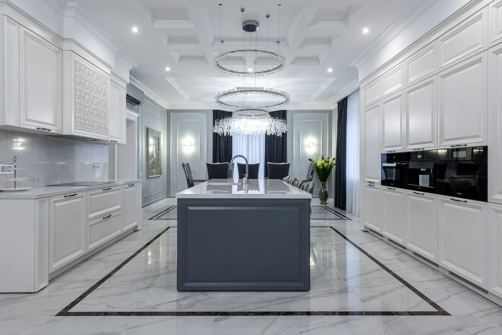 Elegant white kitchen with gray island, marble floors, and chandeliers.
