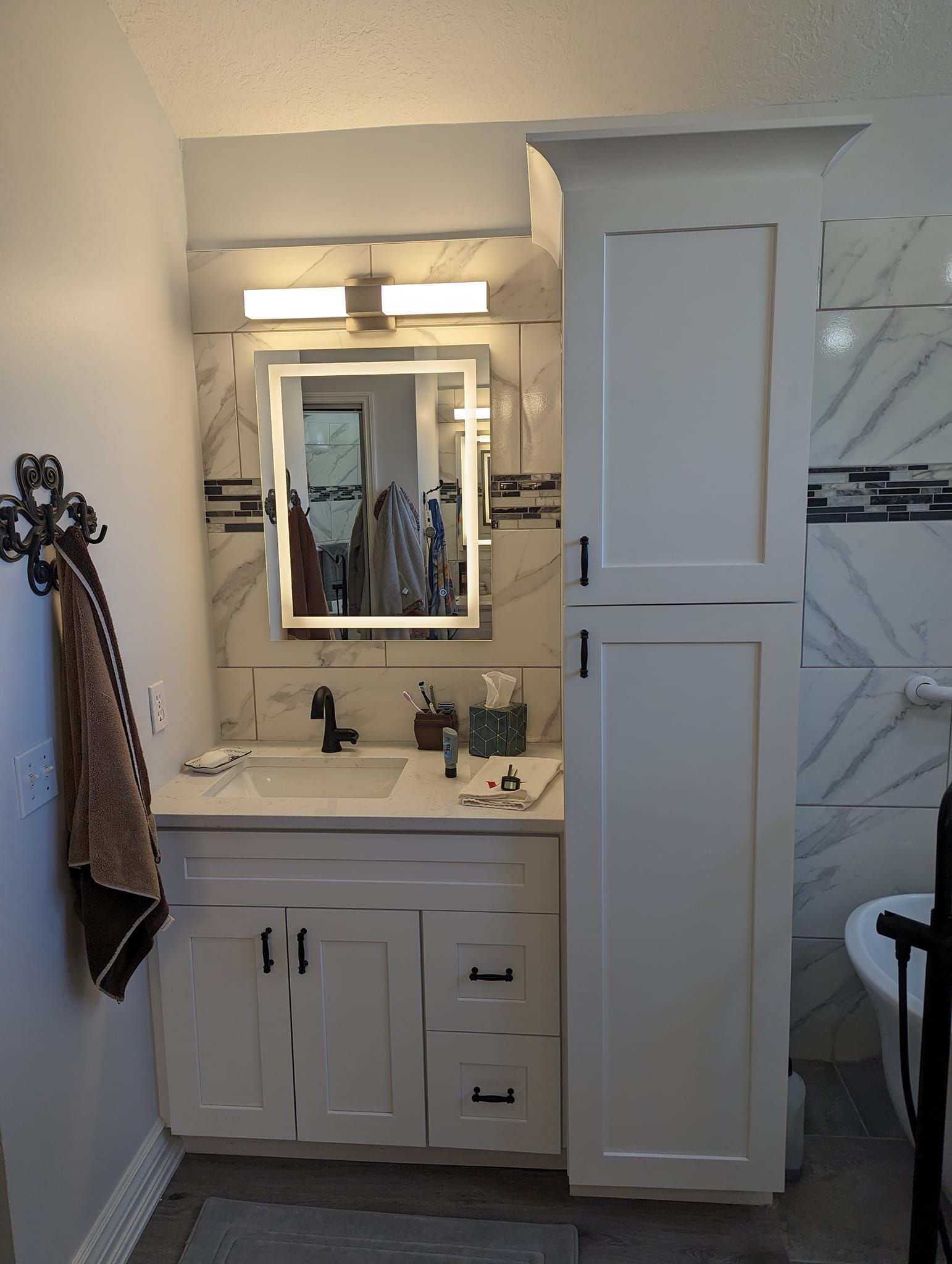Bathroom with white vanity, tall storage cabinet, and marble backsplash. Towel on wall rack.