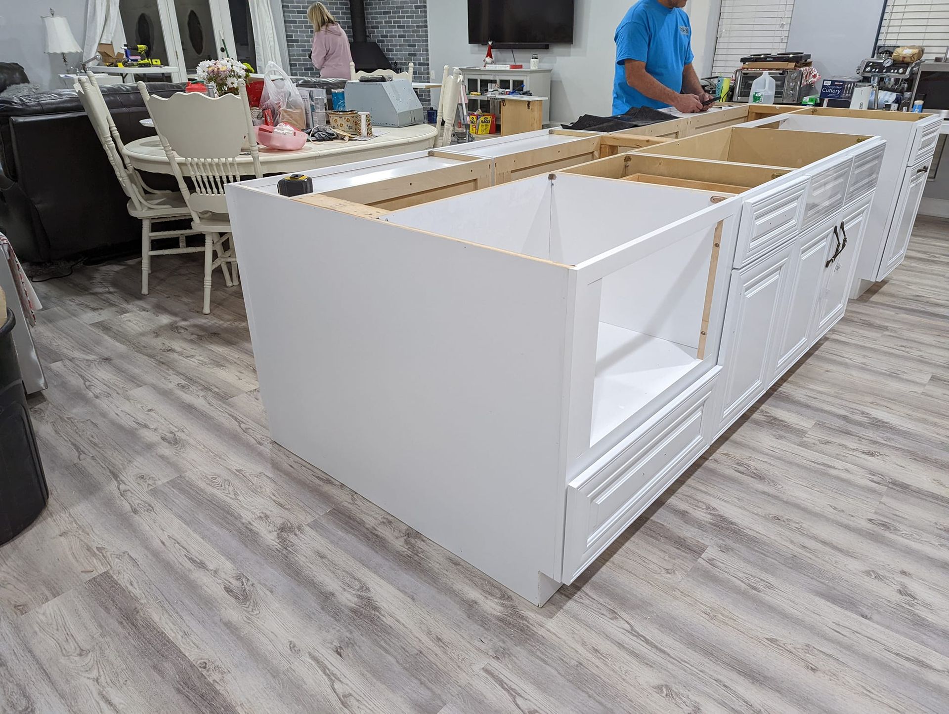White kitchen island cabinets being assembled. A person in blue shirt works on the cabinets, indoors.