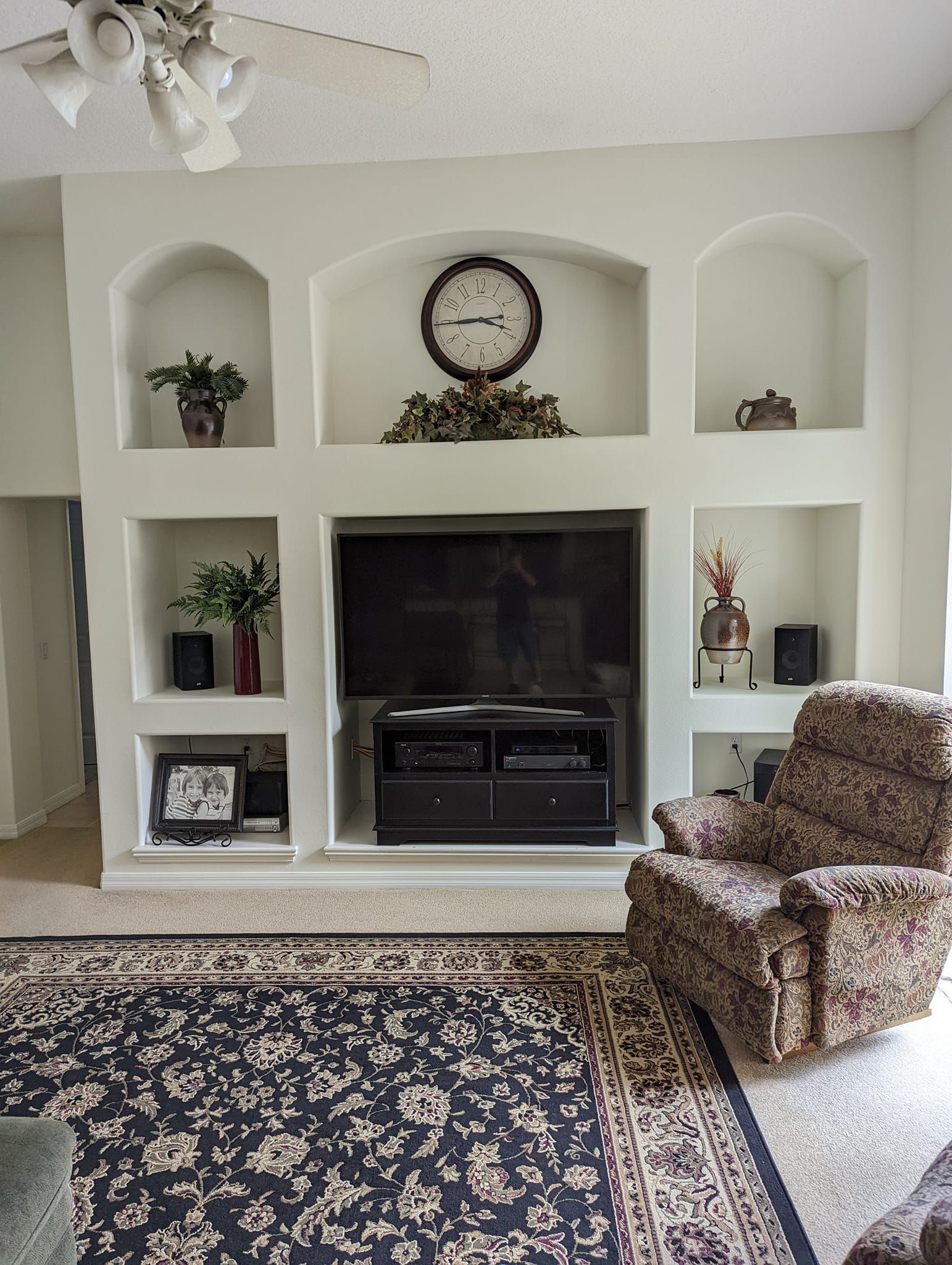 Living room with built-in shelves, TV, clock, and patterned armchair on a rug.