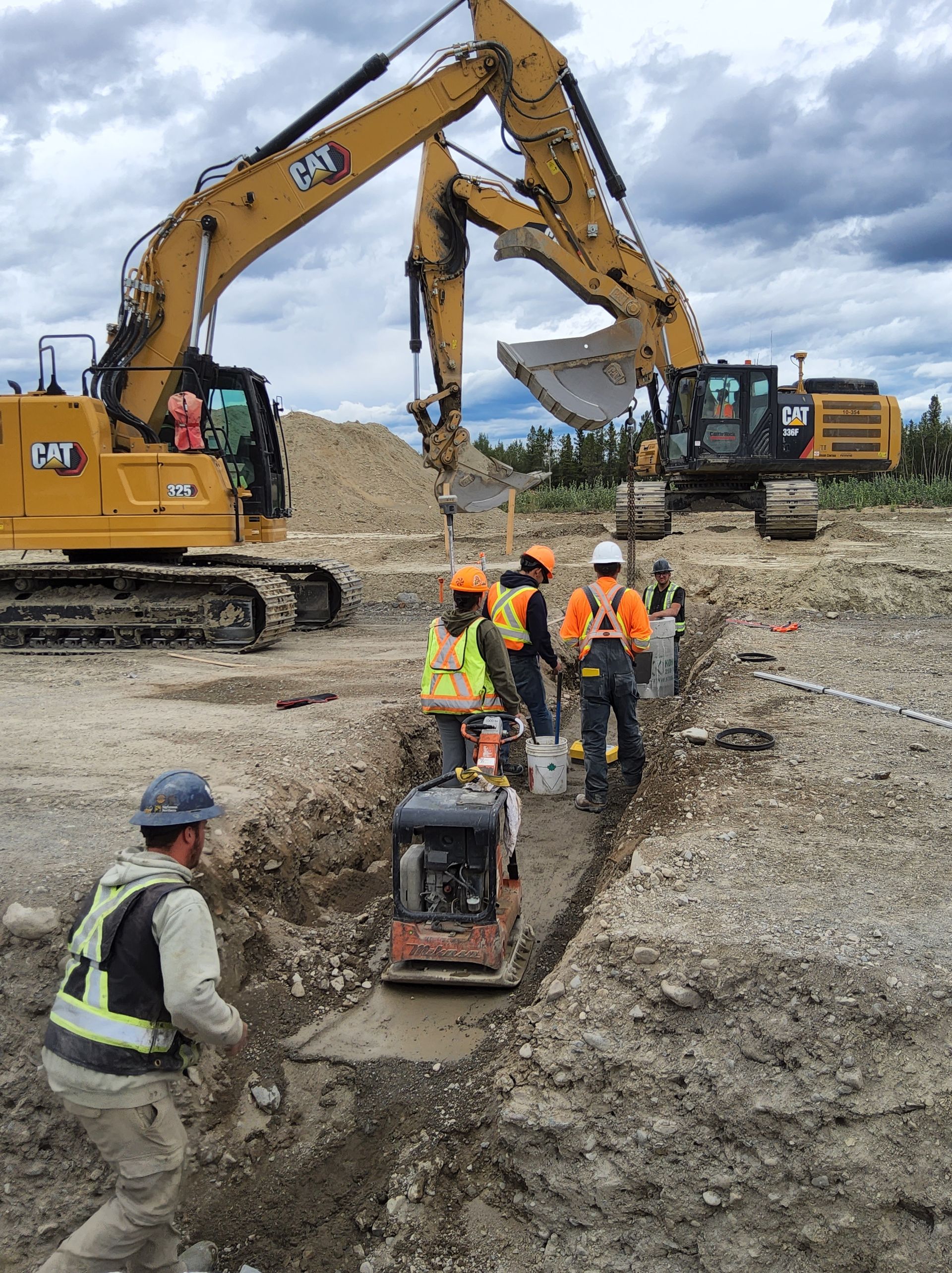 Construction crew compacting a trench, excavators in background. Workers wear safety vests. Outdoors, cloudy sky.