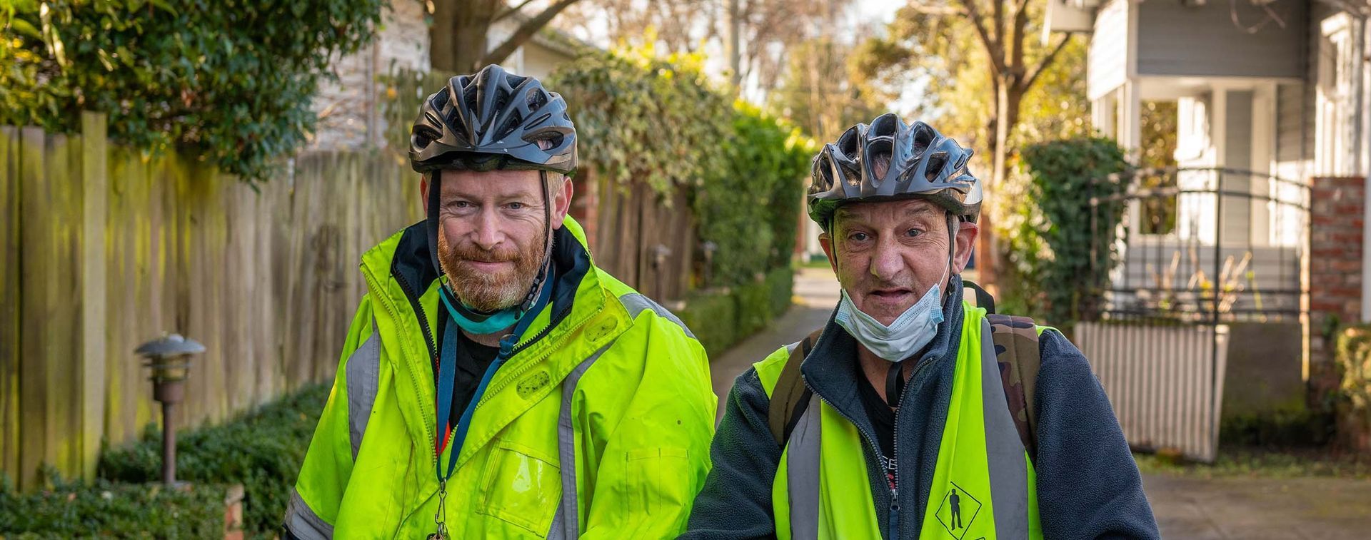 Two men wearing safety vests and helmets are standing next to each other on a sidewalk.