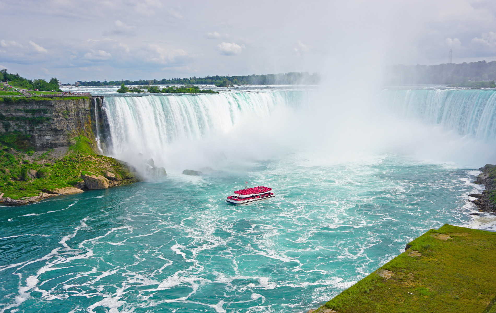 Niagara Falls with boat, turquoise water, white cascading water, and green cliffs under a cloudy sky.