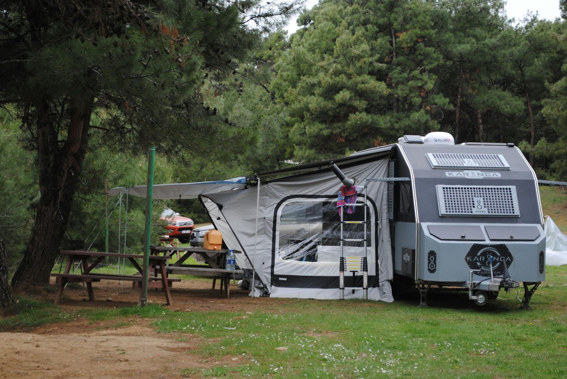 A gray camping trailer with an awning extended, parked near a picnic table in a wooded area.