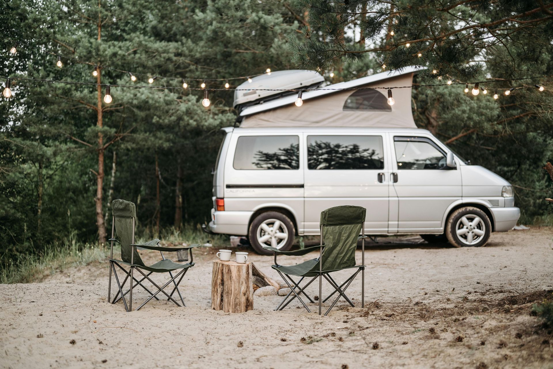 Silver camper van with pop-up roof parked in a wooded area, with folding chairs and string lights.
