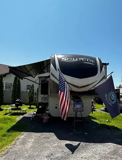 RV trailer with flags parked in front of a house, awning extended, on a sunny day.