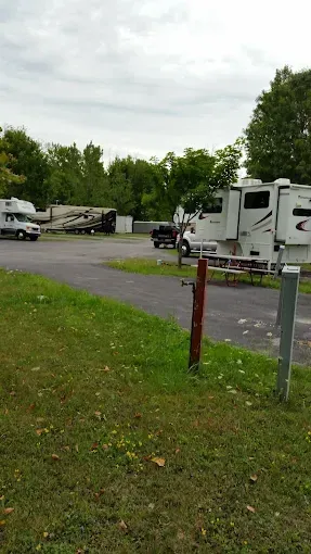 Campground scene with RVs parked on pavement, grass in foreground, trees in background, cloudy sky.