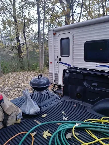 A camping scene with a camper on a truck bed; a grill, firewood, and hose are present.