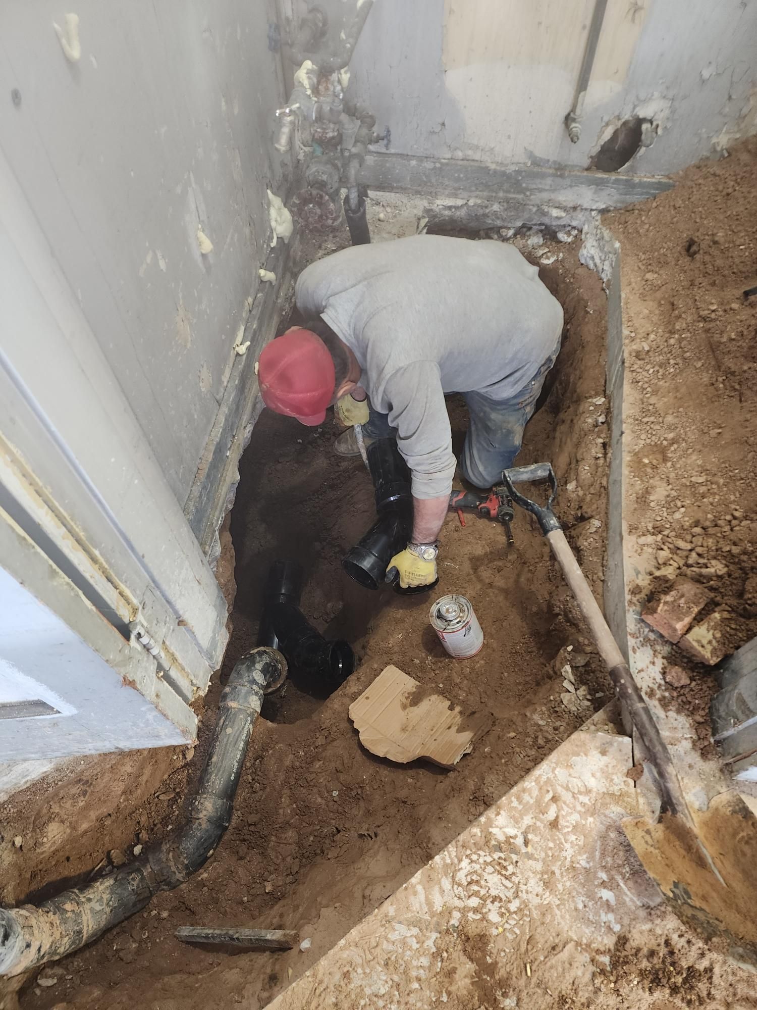 Person in red hat and mask working in a trench, likely plumbing or repair work.