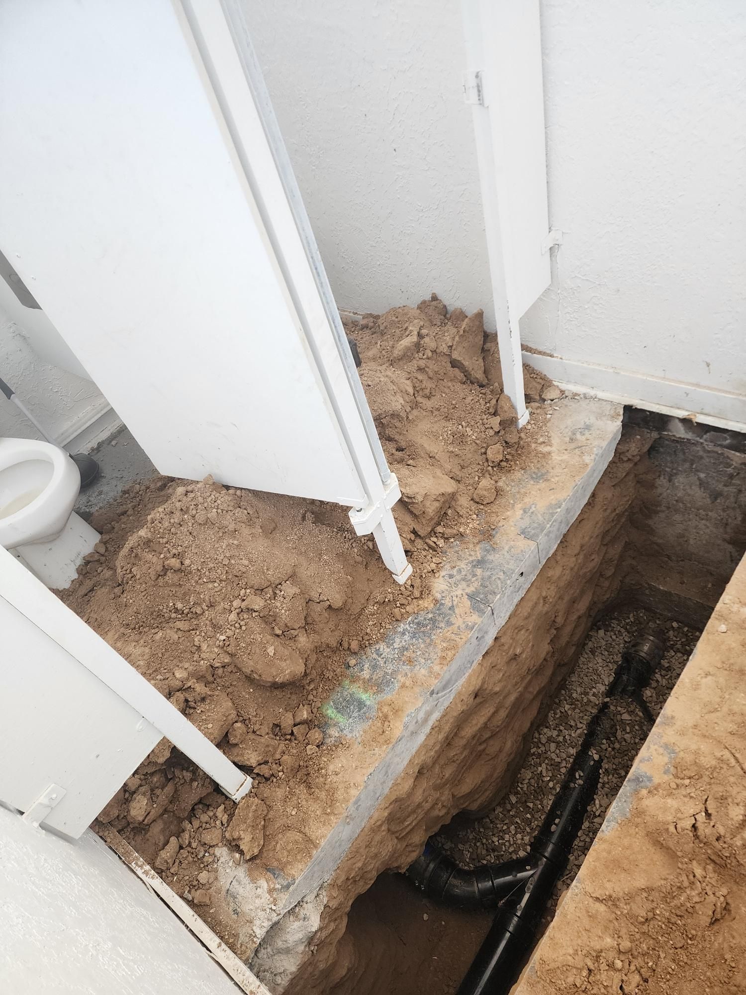 Trench dug in a bathroom floor reveals black plumbing pipes and soil. White walls, a toilet, and a cabinet are visible.