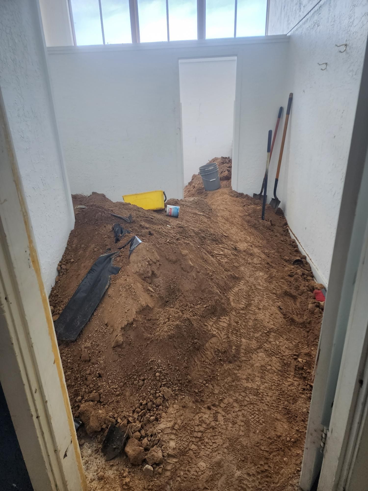 Room filled with dirt; tools lean against the wall. A doorway leads to another room. Overcast lighting.