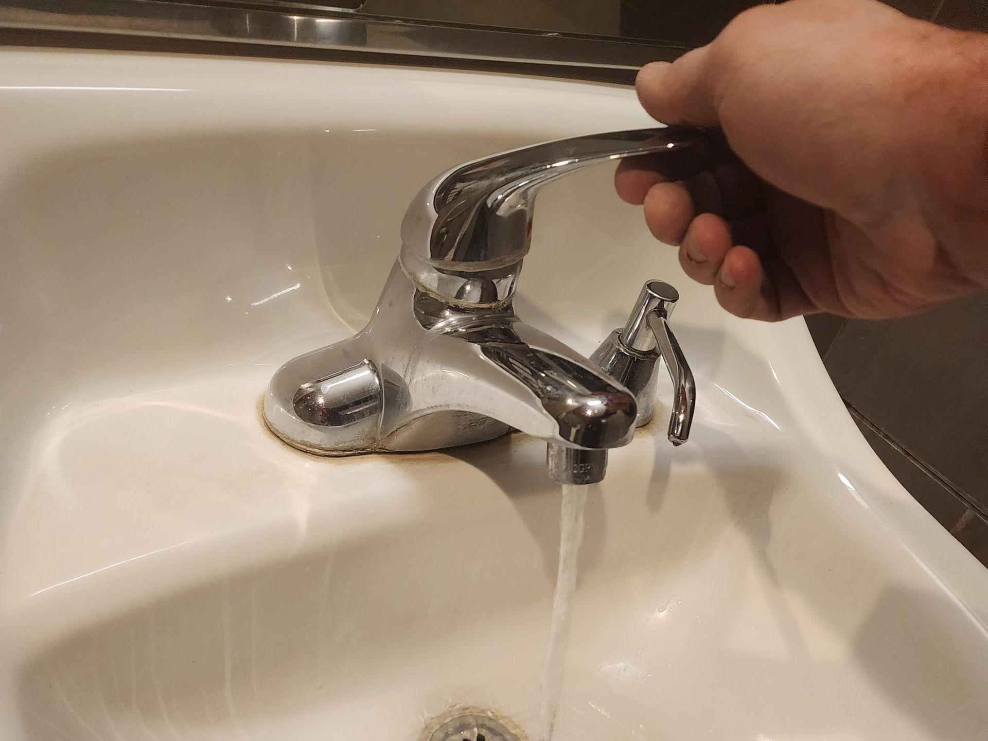 Hand turning on a chrome faucet, water flowing into a white sink.