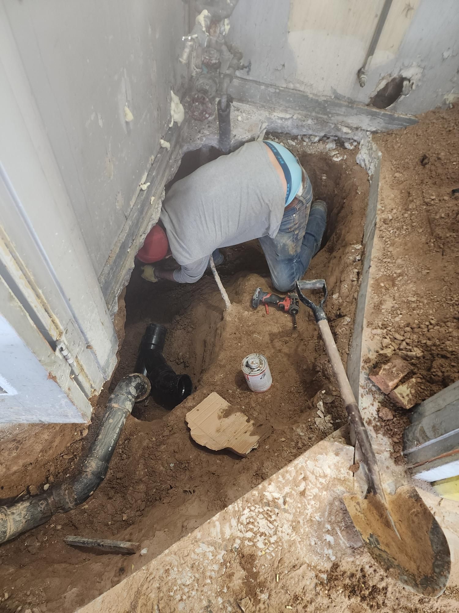 Plumber in a trench, working on plumbing pipes. Earth-toned setting with tools, and a white shirt.