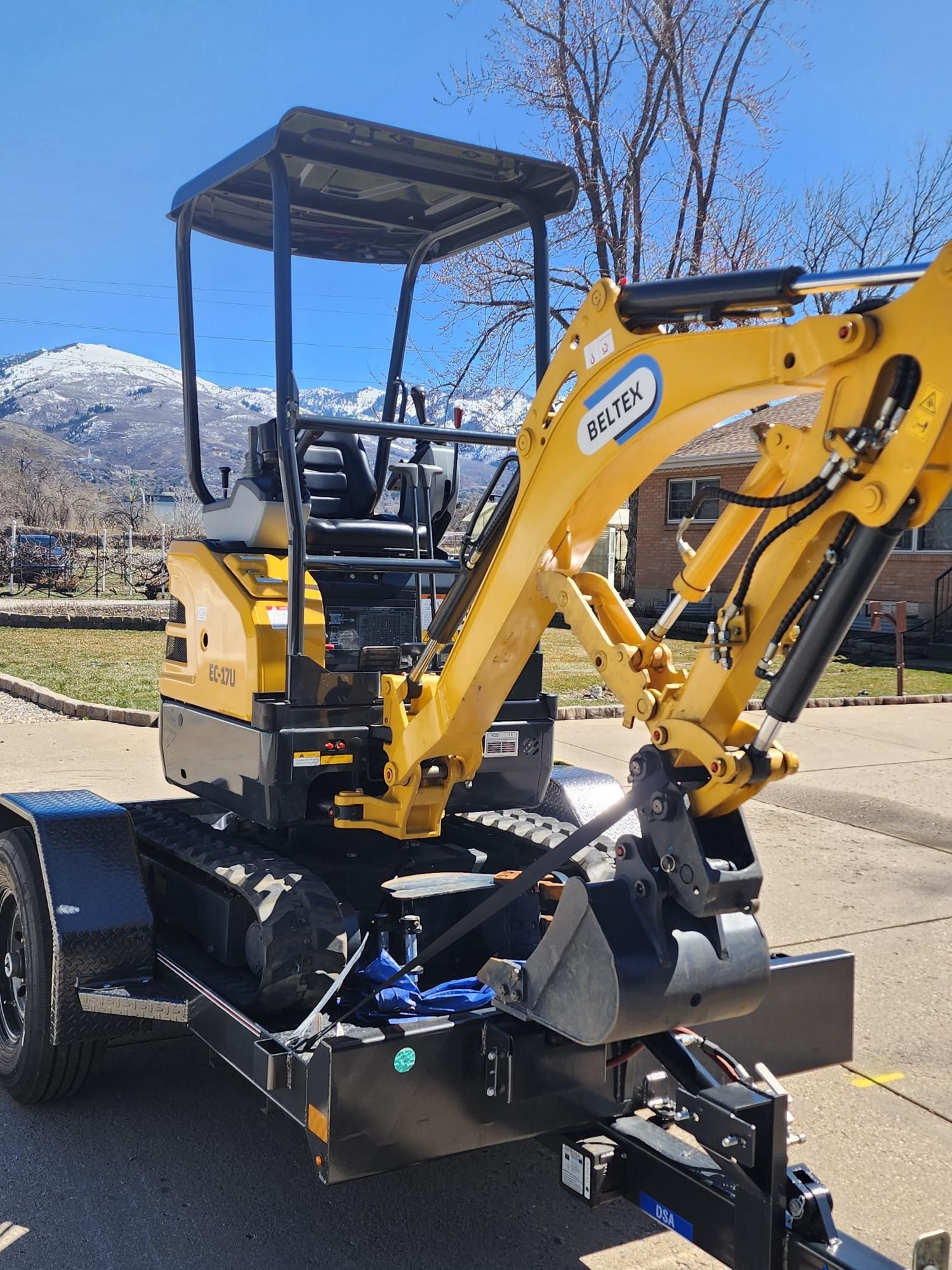 Yellow mini excavator on a black trailer, parked outside on a sunny day with mountains in the background.
