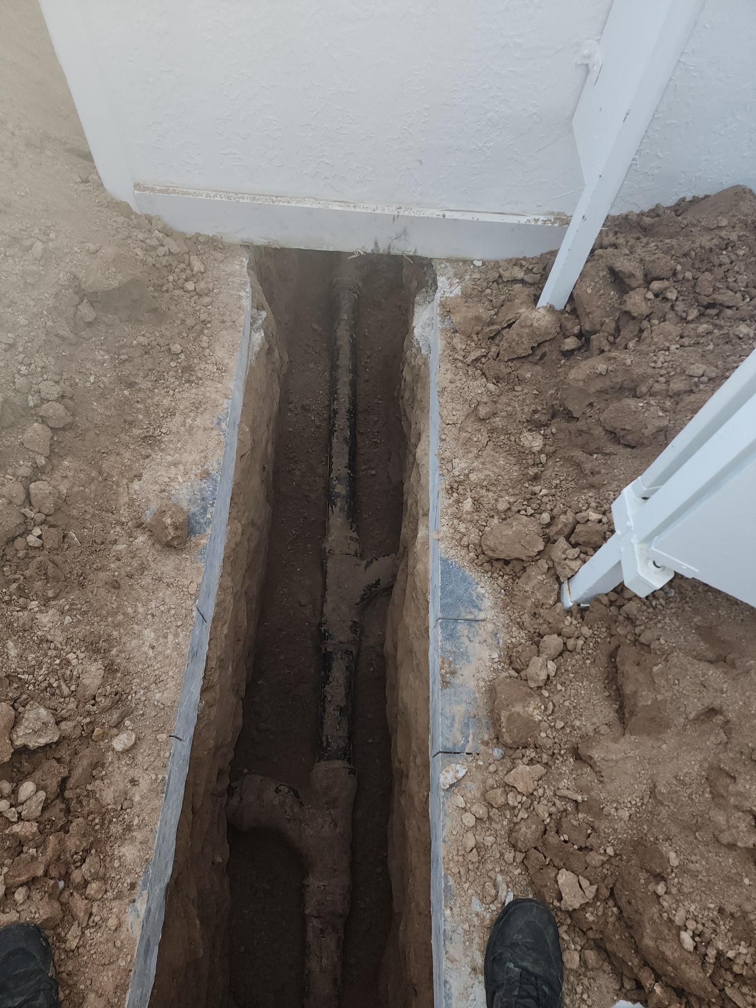 Trench dug alongside a white wall, revealing buried pipes and dirt. A downspout is visible on the right.