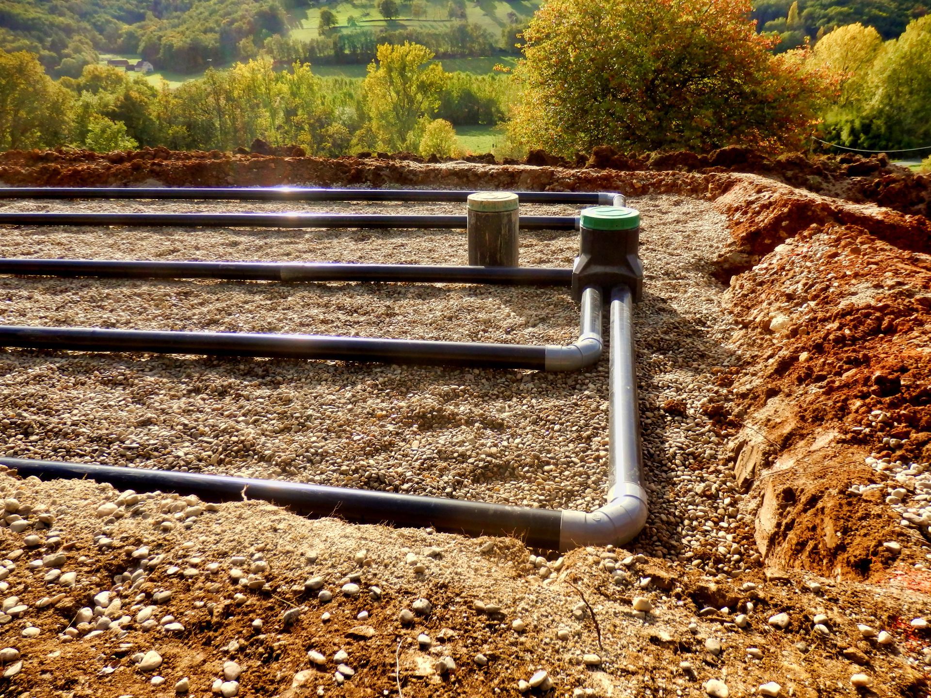 Black pipes in a gravel bed connected to green capped containers, in a rural setting.
