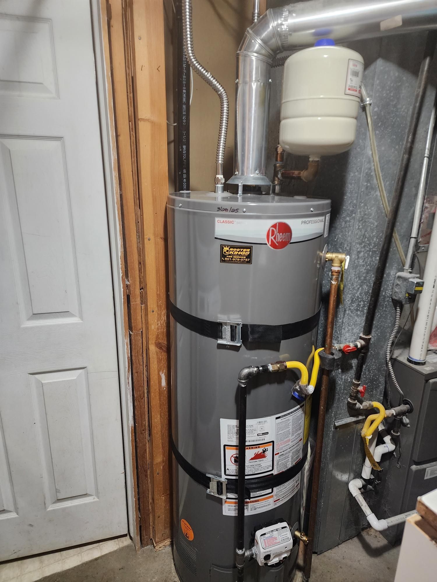 A gray water heater with black straps, vent pipe, and attached components in a utility room.