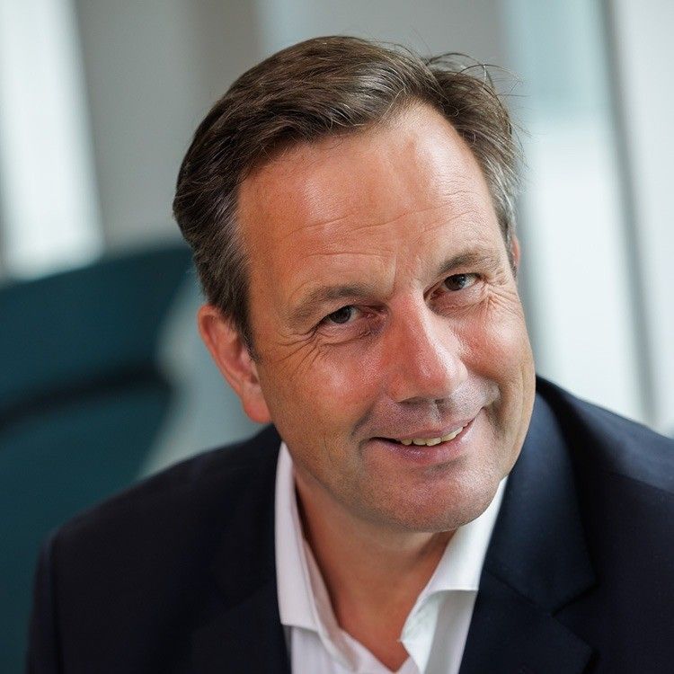 Een man met kort bruin haar glimlacht, gekleed in een donkerblauwe blazer over een wit overhemd, binnenshuis.