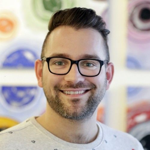 Een man met een bril glimlacht terwijl hij voor een kleurrijk kunstwerk staat.