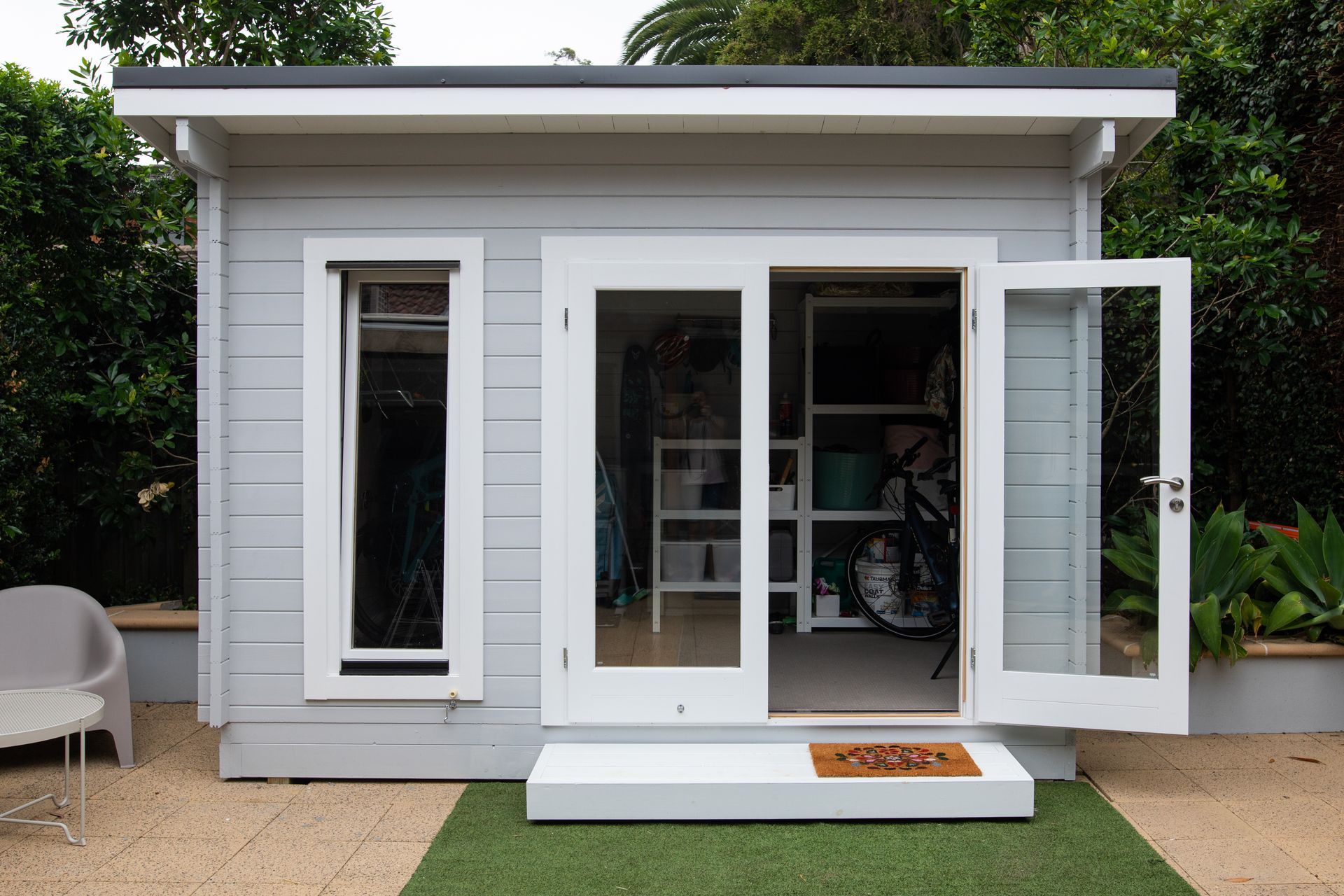 Grey shed with white trim, open double doors, small window, and green doormat.